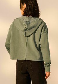 Korte, oversized groene hoodie met een capuchon, gemaakt van zachte stof, met een ruwe zoom en zichtbare stiksels, gecombineerd met zwarte jeans.