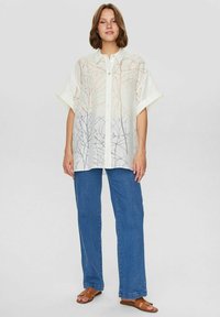 Hvid skjorte med korte ærmer, der har et grenmønster i lysebrune og grå nuancer. Sammen med blå denimbukser og sandaler.