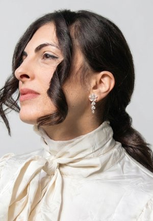 Vrouw met donker golvend haar dat naar achteren is gebonden, draagt een witte blouse met hoge hals en strik, en hanger oorbellen in bloemvorm.