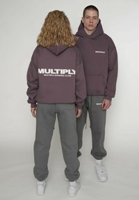 Szara bluza z kapturem z białym logo "MULTIPLY APPAREL CLUB", zestawiona z pasującymi szarymi spodniami dresowymi i białymi tenisówkami, wyróżniająca się casualowym designem.