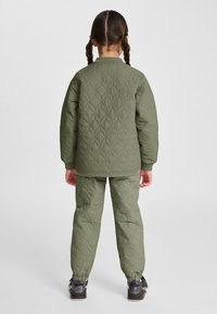 Dziecko o brązowych zaplecionych włosach, ubrane w oliwkowo-zieloną pikowaną kurtkę i spodnie, stojące tyłem na jednolitym tle.