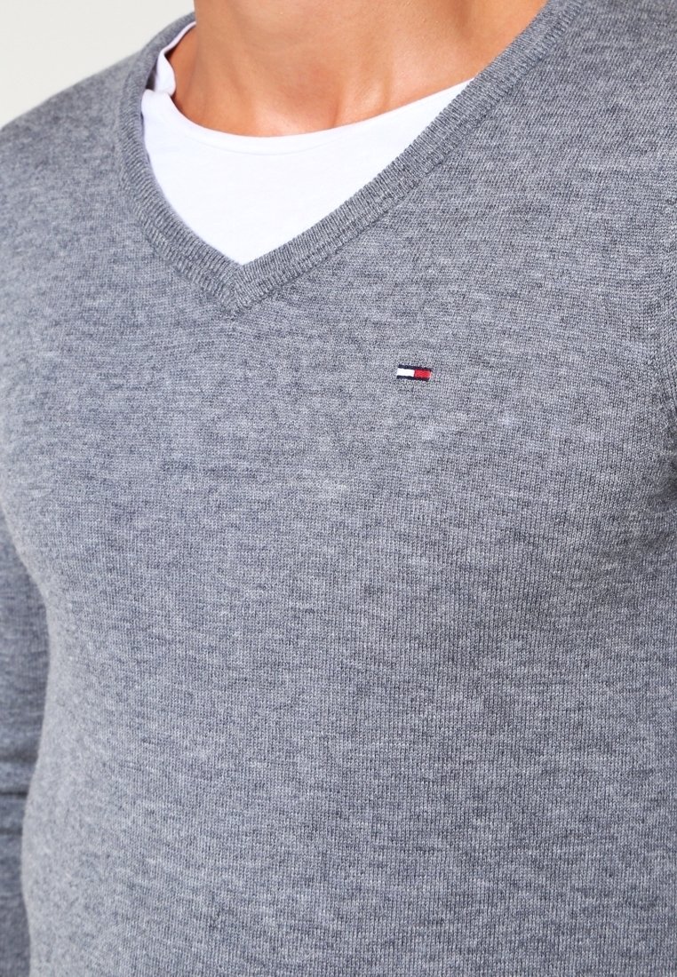 Grauer V-Ausschnitt-Pullover aus weichem Strickmaterial mit subtiler Textur; verfügt über ein kleines Logo in Rot, Weiß und Blau auf der linken Brust.