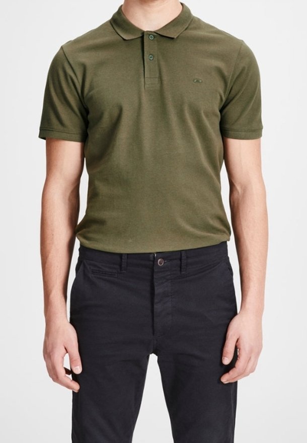 Homme portant un polo à manches courtes vert olive et un pantalon noir, debout devant un fond blanc uni.