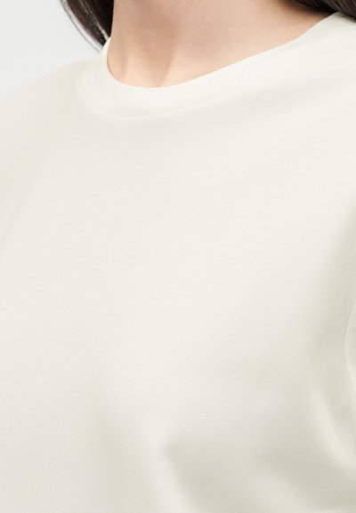 Κοντινό πλάνο ενός ατόμου που φοράει ένα απλό ανοιχτό μπεζ μπλουζάκι με στρογγυλή λαιμόκοψη, εμφανίζοντας τον λαιμό και την περιοχή του άνω στέρνου.