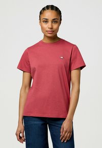 Rode katoenen t-shirt met korte mouwen, ronde halslijn en een klein logo op de borst. Gecombineerd met blauwe jeans.