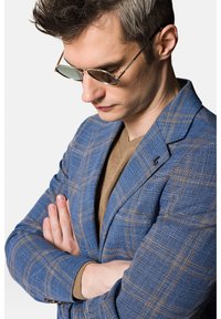 Niebieska marynarka w kratę z teksturowanej tkaniny, zestawiona z beżowym swetrem z dzianiny. Osoba nosi okrągłe, metalowe okulary przeciwsłoneczne.