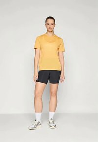 Noor inimene seisab seljas kollane t-särk, mustad jalgrattapüksid, valged sokid ja hallid jooksujalatsid ühtlasel taustal.