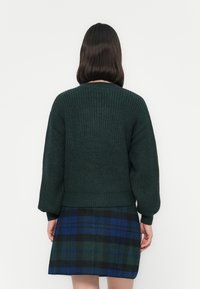 Donkergroene gebreide trui met een ribbeltextuur en brede mouwen. Gepaard met een geruite rok met donkerblauwe, groene en zwarte patronen.