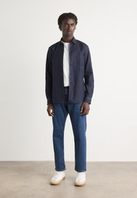 Navy blue button-up shirt with chest pocket, paired with light blue jeans and white sneakers with gum soles, set against a plain background.