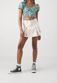 Žena v zeleném květinovém crop topu, béžové plisované mini sukni s kapsami, bílých ponožkách po kotníky a černých vysokých teniskách Converse.