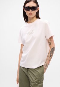 Φωτεινό ροζ βαμβακερό μπλουζάκι με κοντά μανίκια, με κεντημένο λογότυπο "Calvin Klein" σε χρυσό. Μοντέλο που φοράει μαύρα γυαλιά ηλίου και πράσινα παντελόνια.