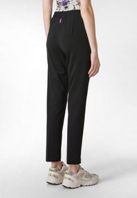 Zwarte, taps toelopende broek met een zachte textuur en een elastische tailleband, met een klein roze label aan de achterkant, gecombineerd met beige sneakers.