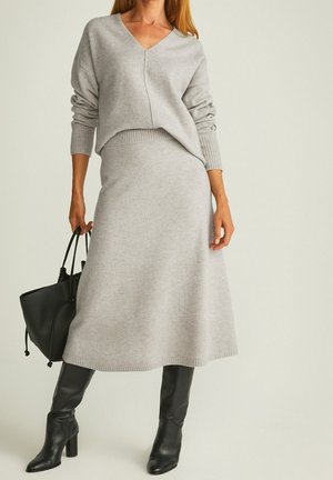 Kvinde i lysegrå strikket sweater og matchende midi nederdel, der holder en sort håndtaske og iført sorte knælange støvler med hæl.