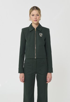 Femme portant une veste zippée vert foncé avec un logo "T" sur la poitrine et un pantalon assorti, debout devant un fond clair uni.
