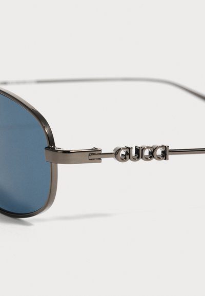 Close-up af metalindfattede blå linse solbriller med tynde metalarme med Gucci-brandlogo i hævede bogstaver.