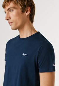 Mladý muž hľadiaci doľava, oblečený v priliehavom tmavomodrom tričku značky Pepe Jeans s logom na hrudi a malou vlajočkovou nášivkou na rukáve.