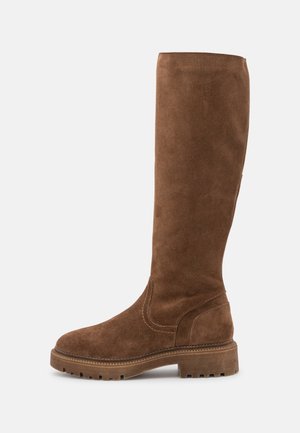 Botte haute en daim marron avec semelle épaisse à crampons et petit talon, dessus lisse avec coutures visibles le long de la semelle et de la zone de la cheville.