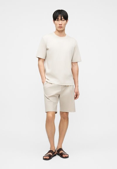 Νέος άνδρας όρθιος, φοράει μπεζ κοντομάνικο μπλουζάκι και ταιριαστό σορτς με σκούρα καφέ σανδάλια ανοιχτά στα δάχτυλα, μπροστά σε απλό φωτισμένο φόντο.