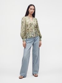 Femme portant un chemisier transparent à fleurs vert clair avec de longues manches bouffantes et un jean large bleu clair, debout devant un fond blanc.
