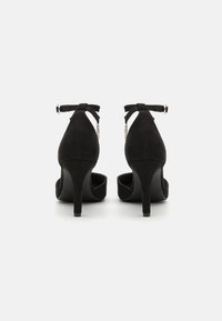 Scarpe con tacco alto in suede nero con cinturini alla caviglia, punta aperta e una texture liscia. Gli accessori in metallo argento sul cinturino aggiungono dettagli.