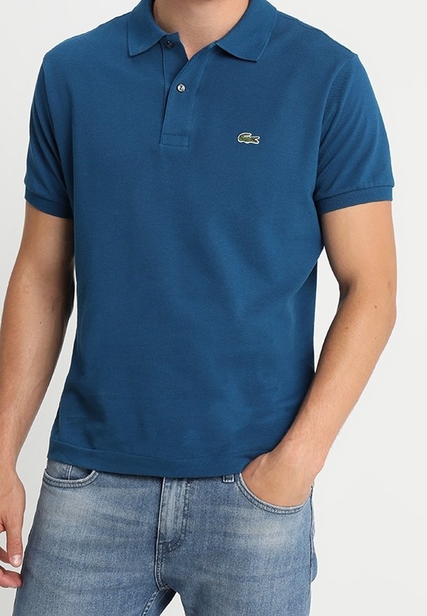 Homme portant un polo bleu à manches courtes avec le logo Lacoste et un jean denim bleu clair, une main dans la poche, sur fond blanc uni.