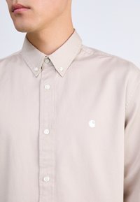 Camisa beige de botones con un cuello clásico, botones blancos y un pequeño logo blanco en el lado izquierdo del pecho. Textura de algodón suave.