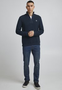 Marineblauer Reißverschluss-Pullover mit strukturierten Streifen, schwarze Jeans und schwarze Sneakers. Verfügt über ein kleines Logo auf der Brust sowie Rippbündchen und einen gerippten Saum.