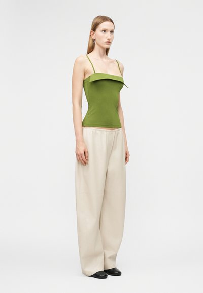 Γυναίκα στέκεται φορώντας πράσινο αμάνικο τοπ με λεπτές τιράντες και μπεζ παντελόνι με φαρδιά πόδια, μπροστά σε απλό φωτεινό φόντο.