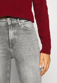 Jeans de mezclilla gris de cintura alta, con cierre de botón, cinco bolsillos y una textura suave y ligeramente deslavada. Combinados con un suéter de punto rojo.