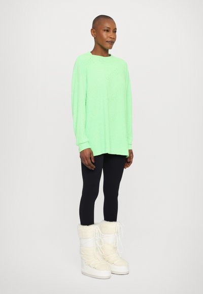Maglietta verde brillante a maniche lunghe con design testurizzato "SKGLTZ", abbinata a leggings neri e stivali invernali foderati in pelliccia bianca.