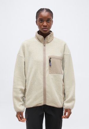 Jonge persoon met gevlochten haar draagt een beige fleecejack met rits en borstzak, staand tegen een effen witte achtergrond.