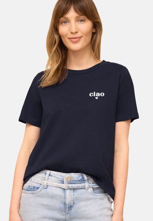 Kobieta w granatowym t-shircie z napisem "ciao" i małym serduszkiem na piersi, w połączeniu z jasnoniebieskimi dżinsami.