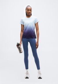 Γυναίκα με μπλουζάκι προπόνησης σε μπλε διαβάθμιση χρωμάτων, μπλε κολάν και λευκά αθλητικά παπούτσια, κρατώντας ένα μπουκάλι νερού Puma, στέκεται μπροστά σε απλό φόντο.