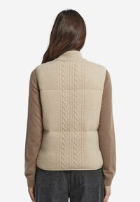 Beige stickad väst med ett kabelmönster på baksidan. Framsidan har bruna ärmar, en hög krage och en strukturerad yta.