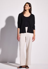 Cardigan nero con una vestibilità rilassata e apertura frontale, abbinato a pantaloni in lino beige chiaro. Il modello indossa sandali piatti neri. Design minimalista.