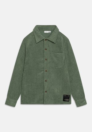 Camisa de pana verde con mangas largas, cierre de botones al frente y un bolsillo en el pecho. Presenta una superficie texturizada y una etiqueta de la marca cerca del dobladillo.