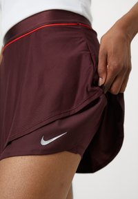 Burgundi színű sportnadrág bordázott textúrával, piros díszcsíkkal és ezüst Nike logóval az alsó szegélyen.