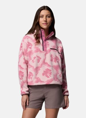 Kvinde iført en pink og hvid mønstret fleece-pullover med trykknapper og grå shorts, stående mod en hvid baggrund.