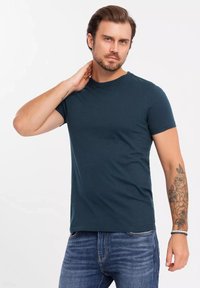 Tmavě modrá tričko s kulatým výstřihem z měkké tkaniny, krátké rukávy a přiléhavý střih. Na předloktí modelu je viditelný tetování.