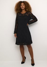 Schwarzes Spitzenkleid mit langen Ärmeln und einem gewellten Ausschnitt, versehen mit einem strukturierten Blumenmuster und einem knielangen Saum. Getragen mit schwarzen High Heels.