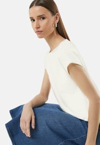 Haut blanc texturé à manches courtes, associé à une jupe en denim foncé. La silhouette a des cheveux longs et raides et porte des boucles d'oreilles simples en anneau.