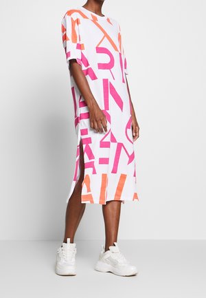 Person trägt ein weißes Midikleid mit knallpinken und orangenen abstrakten Buchstabenmustern, seitlichen Schlitzen und weißen, klobigen Sneakers.