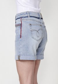 Shorts en denim bleu clair avec des ourlets retroussés, arborant des coutures décoratives en rouge et en bleu le long de la taille et des poches arrière.