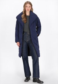 Marineblå quiltet lang frakke med oversized krave, der har trykknapper, tekstureret stof og sideslidser. Kombineret med mørke jeans og ankelstøvler.