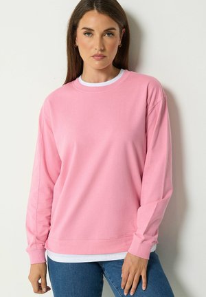 Donna con i capelli castani lunghi che indossa una felpa rosa a maniche lunghe sopra una camicia bianca e jeans blu, in piedi contro un muro bianco.