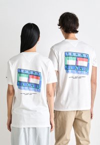Las camisetas blancas de algodón presentan un diseño gráfico colorido en la parte posterior con el texto "TOMMY JEANS" y nombres de ciudades en rectángulos verdes y azules.