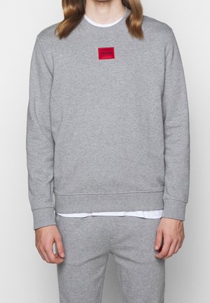 Person, der einen hellgrauen Pullover und passende Jogginghosen mit einem roten "HUGO"-Logo-Patch auf der Brust trägt.