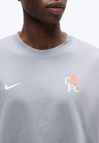 Κοντινό πλάνο ενός ατόμου που φοράει ανοιχτό γκρι πουκάμισο με λευκό λογότυπο της Nike και ένα πορτοκαλί λιοντάρι πάνω από τα γράμματα «CFC» στο στήθος.