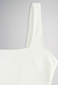 Haut en tissu blanc côtelé avec un décolleté carré et une large bretelle sur un fond neutre.