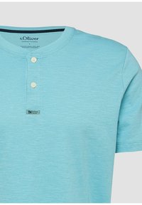 Chemise henley bleu clair à manches courtes avec deux boutons blancs et petite étiquette "Elevated Casual" sur la couture de la poitrine.